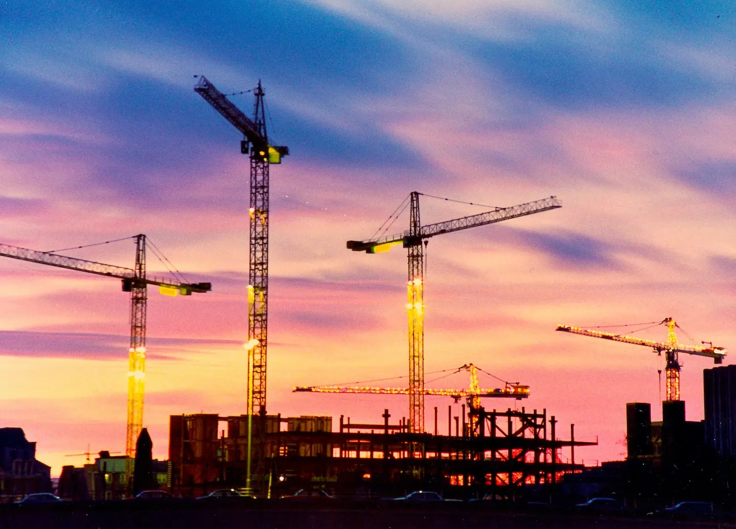 Five cranes on construction site during sunset.
