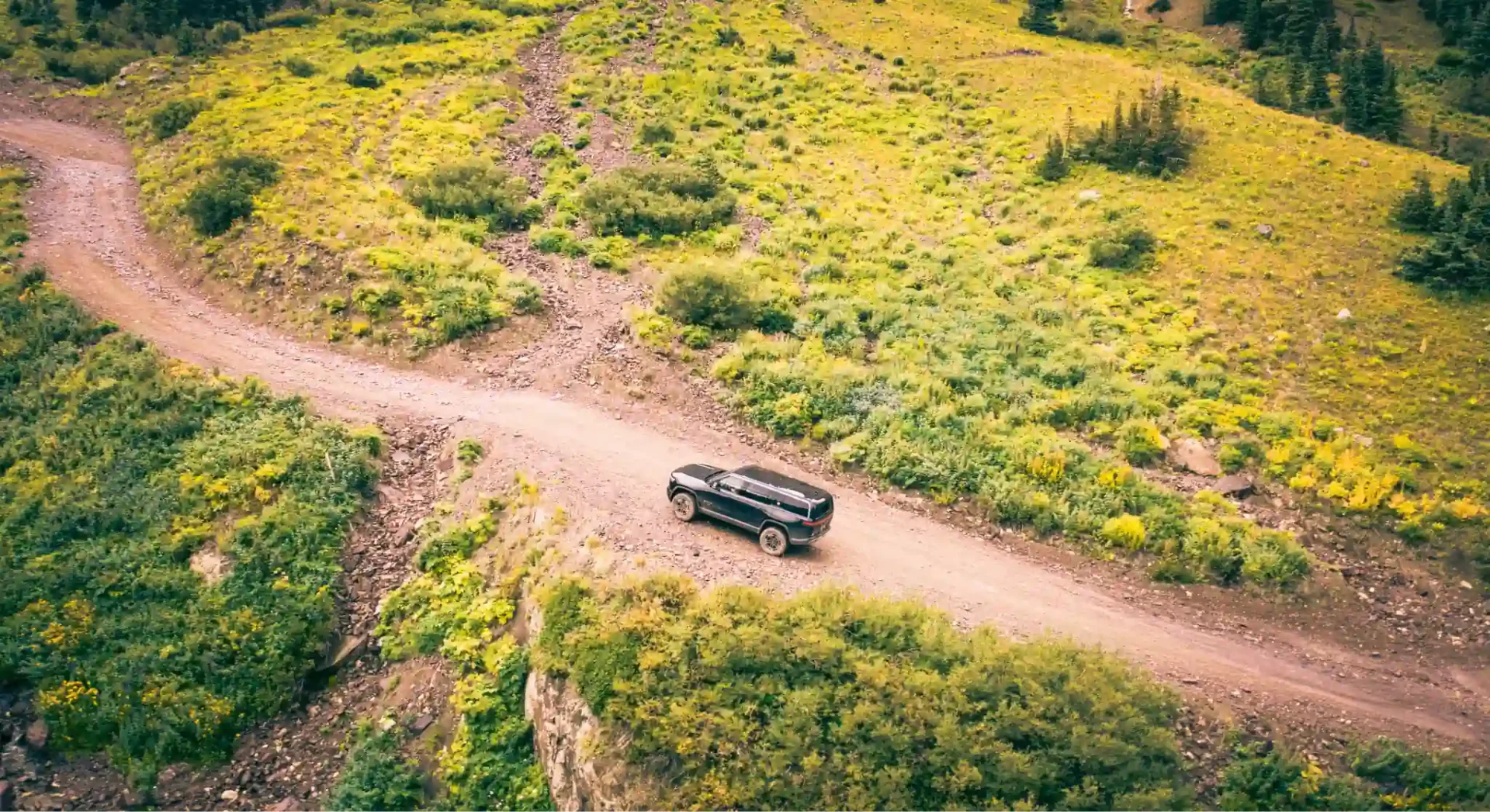 Rivian SUV traveling along dirt path in hilly terrain.